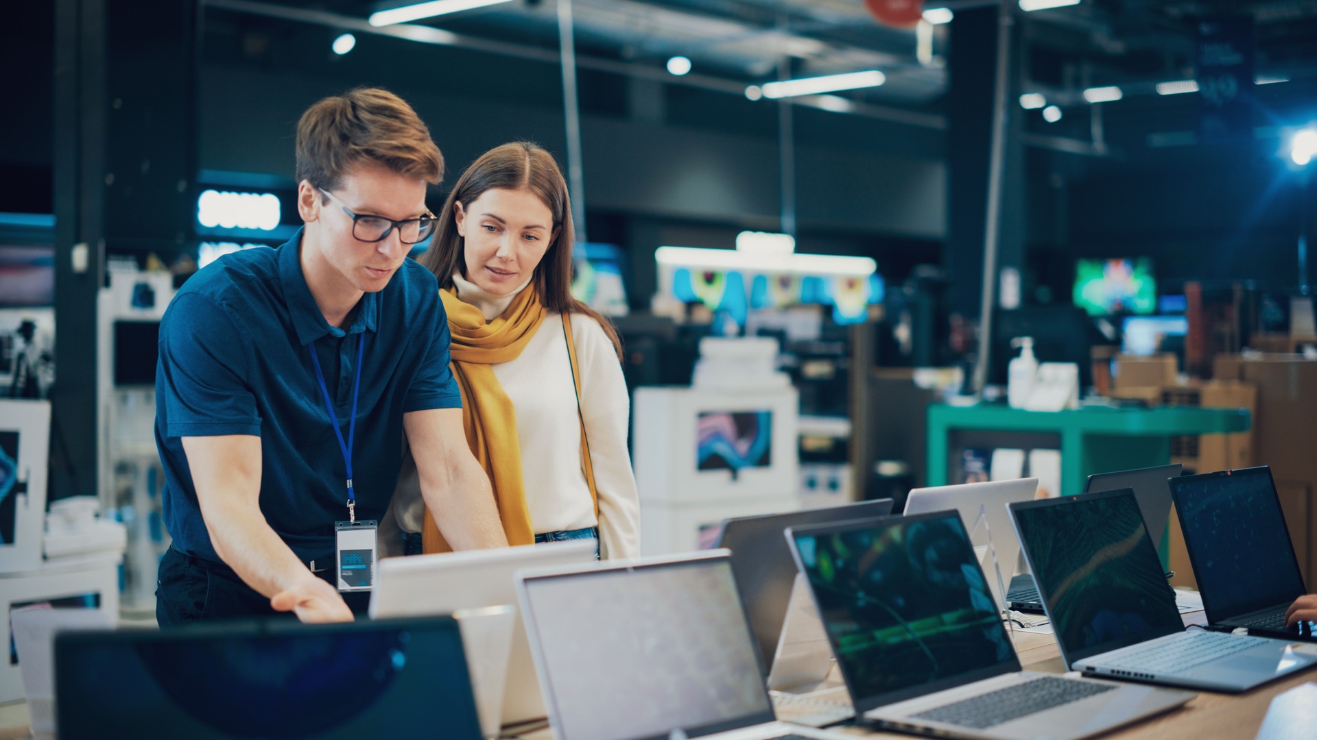 Attractive Woman Discusses Laptop Options with Home Electronics Store Sales Associate. Young Shopper Examines Modern Computer Choices with the Help of a Sales Expert in a Showroom.